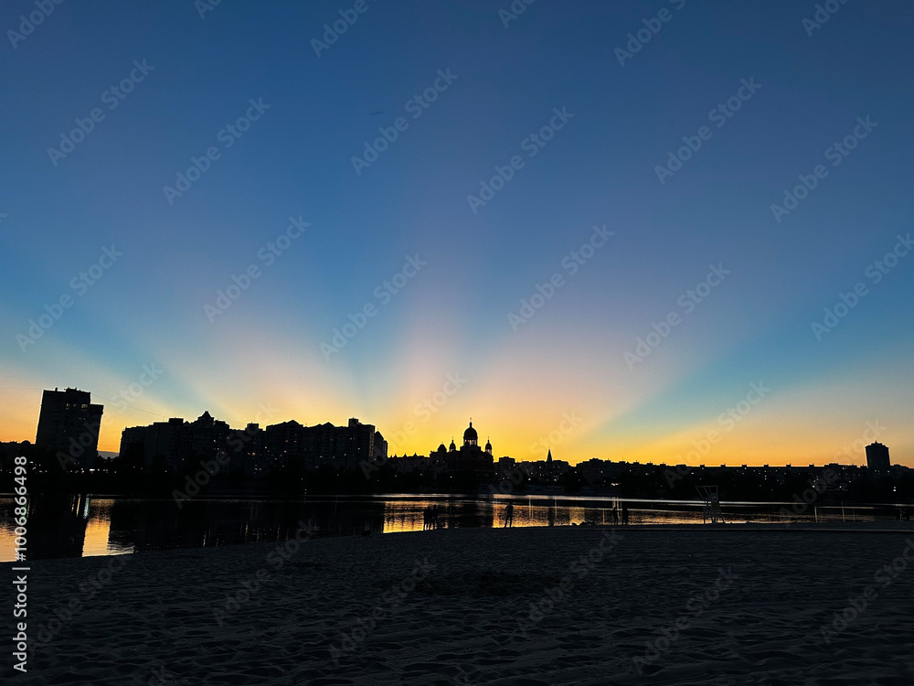 shows a stunning sunset over a city skyline, with rays of light spreading across the sky. The silhouettes of buildings and a church with domes are reflected in the calm water below. shows a stunning sunset over a city skyline, with rays of light spreading across the sky. The silhouettes of buildings and a church with domes are reflected in the calm water below.