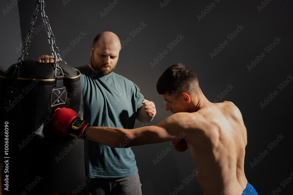 A strict trainer watches his teenager male kickboxer student practice a punch on a punching bag A strict trainer watches his teenager male kickboxer student practice a punch on a punching bag