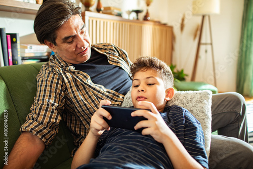 Father and son relaxing on couch watching video on smartphone together photo