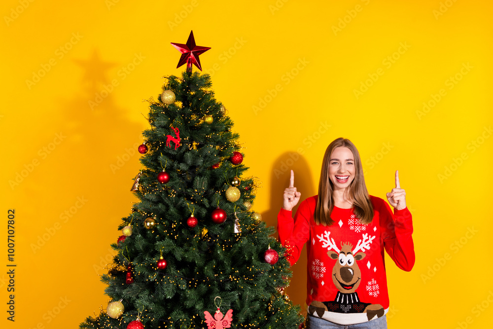 Portrait of nice young girl point fingers up empty space decorated tree wear festive new year sweater isolated on yellow color background Portrait of nice young girl point fingers up empty space decorated tree wear festive new year sweater isolated on yellow color background