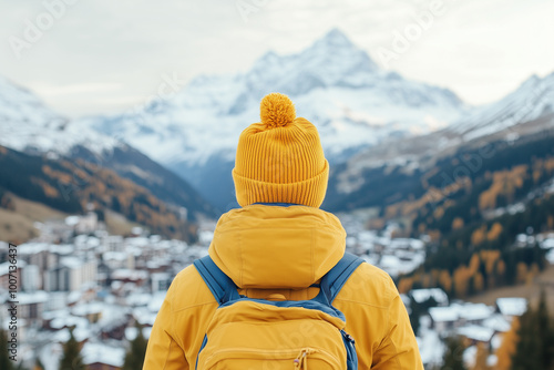 Back view of a traveler wearing a yellow beanie and backpack overlooking a snowy mountain village during autumn