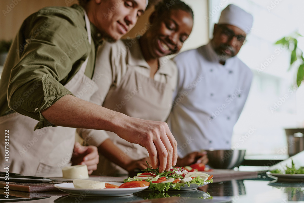 Medium close up of young sous chef adding condiment into caprese appetizer while female cook and chef looking at him Medium close up of young sous chef adding condiment into caprese appetizer while female cook and chef looking at him