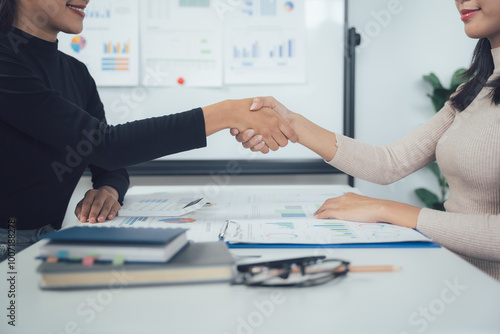 Businesswomen Handshake: Two professional women shake hands across a table in a modern office, symbolizing a successful partnership, agreement, or negotiation. The image conveys trust, collaboration.