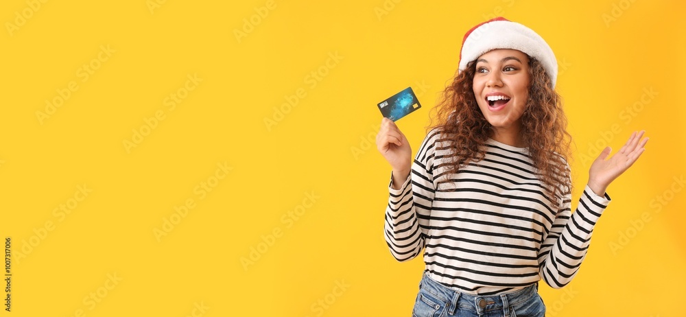 Happy African-American woman in Santa hat holding credit card on yellow background with space for text Happy African-American woman in Santa hat holding credit card on yellow background with space for text