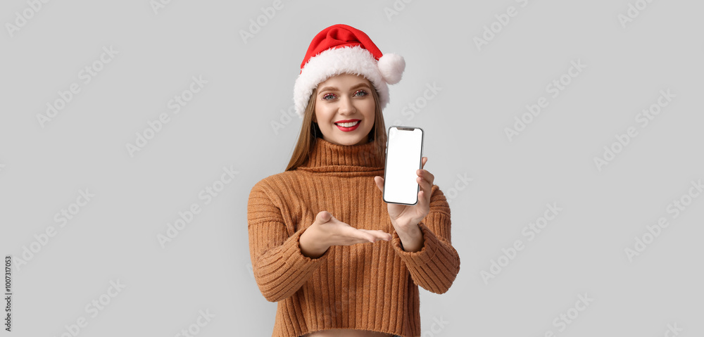 Fototapeta premium Beautiful young woman in Santa hat and with mobile phone on grey background