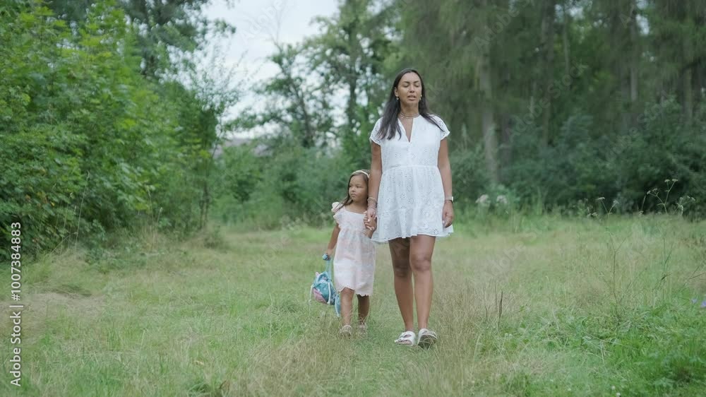 A pregnant woman and her young daughter, both dressed in white, walk hand-in-hand through a grassy park. The scene captures a serene moment of family bonding in nature, surrounded by trees. A pregnant woman and her young daughter, both dressed in white, walk hand-in-hand through a grassy park. The scene captures a serene moment of family bonding in nature, surrounded by trees.