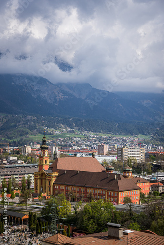 Panoramic view of Innsbruck, Tyrol, Austria, featuring the Stiftskirche (Abbey Church) with its beautiful Baroque design, set against the stunning backdrop of the Nordkette mountain range and vibrant 