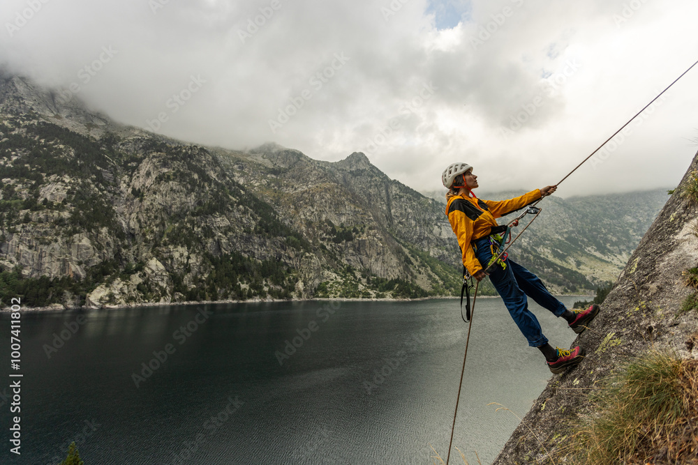 Obraz premium A man is climbing a rock face with a rope attached to him. The scene is set against a backdrop of mountains and a lake. The man is wearing a yellow jacket and he is enjoying the challenge of the climb