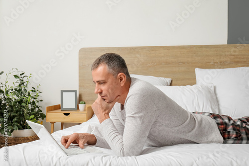 Morning of handsome mature man in stylish pajamas with laptop lying on bed at home