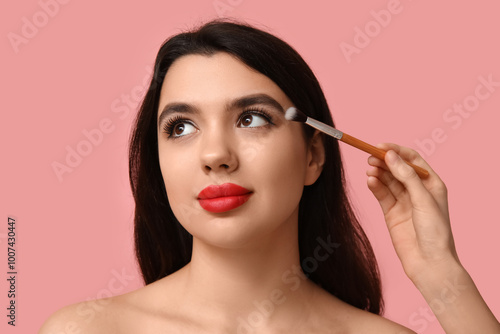 Female makeup artist working with young woman on pink background