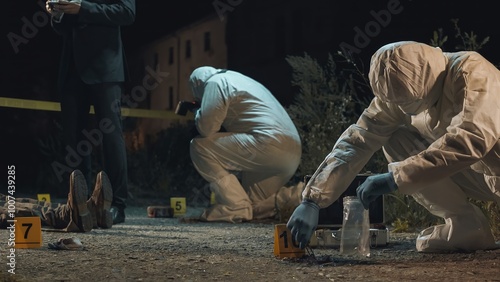 Detective and forensic scientists working hard at crime scene near headless body. Collection and photography of evidence, measurements, examination of the victim's body. Night scene.
