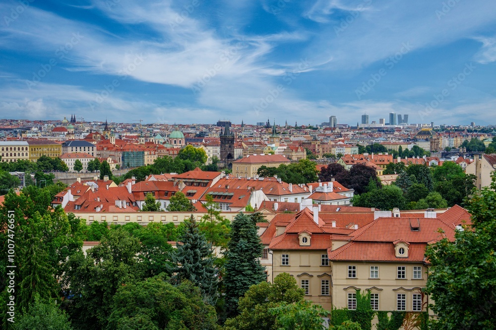 Obraz premium This image showcases an aerial view of Prague, featuring its iconic red-roofed buildings and lush greenery. The blue sky and white clouds enhance the beauty of this stunning cityscape.