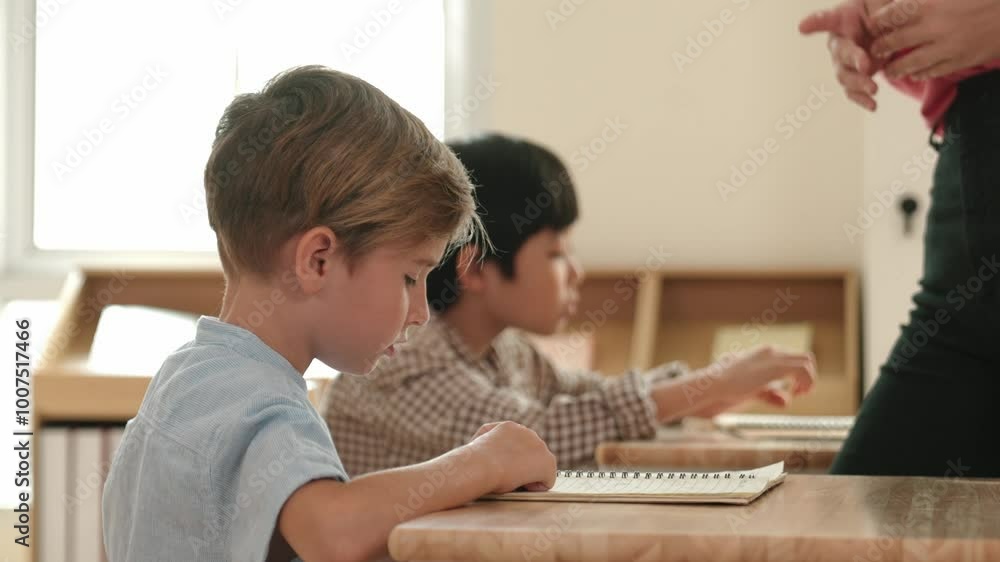 Side view of caucasian elementary student looking a note and answer question while sitting in front of blackboard. Young teacher explain classwork or test while children attend the class. Pedagogy. Side view of caucasian elementary student looking a note and answer question while sitting in front of blackboard. Young teacher explain classwork or test while children attend the class. Pedagogy.