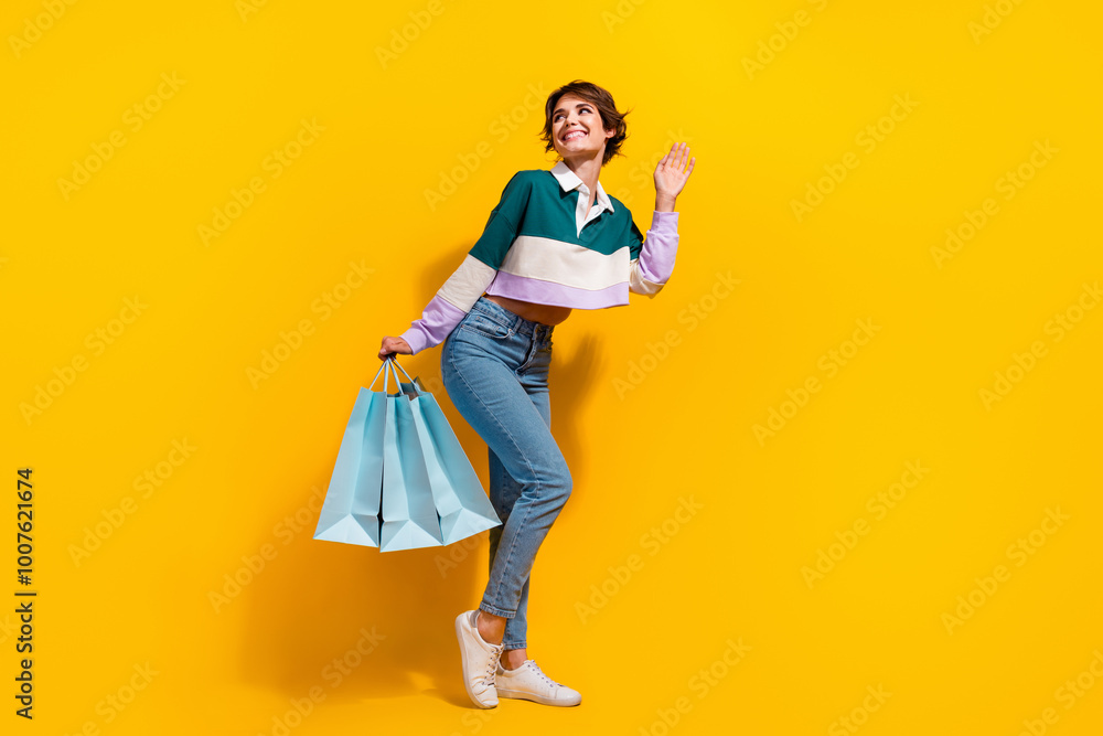 Full length photo of friendly cute woman wear polo shirt waving hi holding bargains looking empty space isolated yellow color background Full length photo of friendly cute woman wear polo shirt waving hi holding bargains looking empty space isolated yellow color background