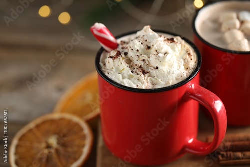 Tasty hot cocoa drink with candy cane and whipped cream in red mugs on table, closeup