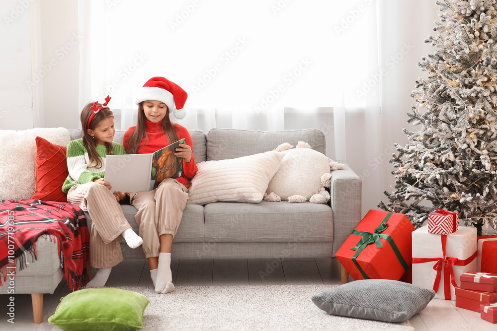 Fototapeta premium Cute happy little girls with book reading Christmas story on sofa at home