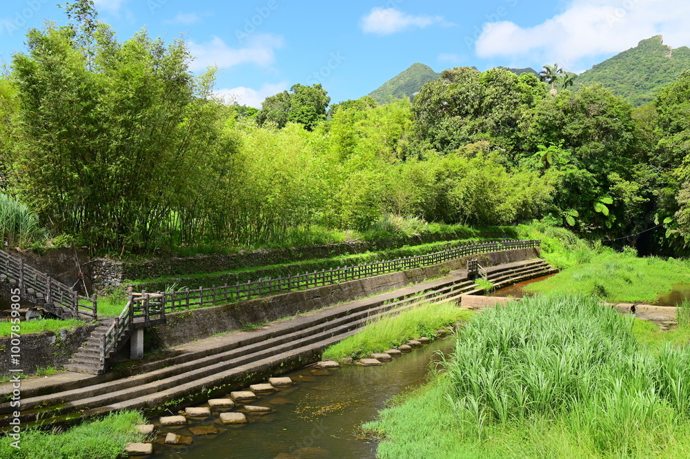 On a bright day with blue skies and white clouds, a concrete-reinforced riverbank complements the lush greenery along a scenic hiking trail, showcasing a balance of nature and engineering. On a bright day with blue skies and white clouds, a concrete-reinforced riverbank complements the lush greenery along a scenic hiking trail, showcasing a balance of nature and engineering.