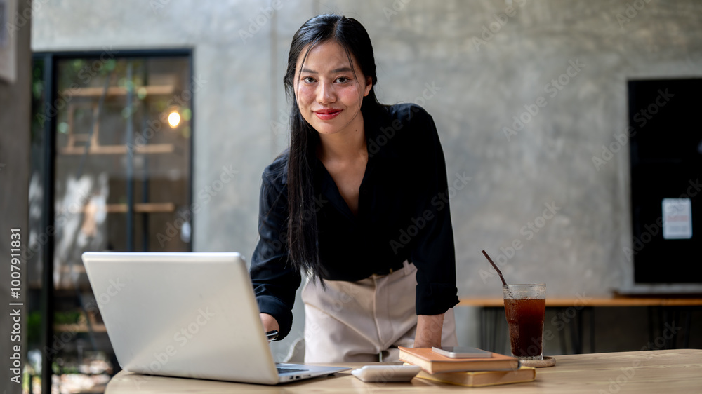 An attractive, confident Asian businesswoman is smiling at the camera while leaning on her desk. An attractive, confident Asian businesswoman is smiling at the camera while leaning on her desk.