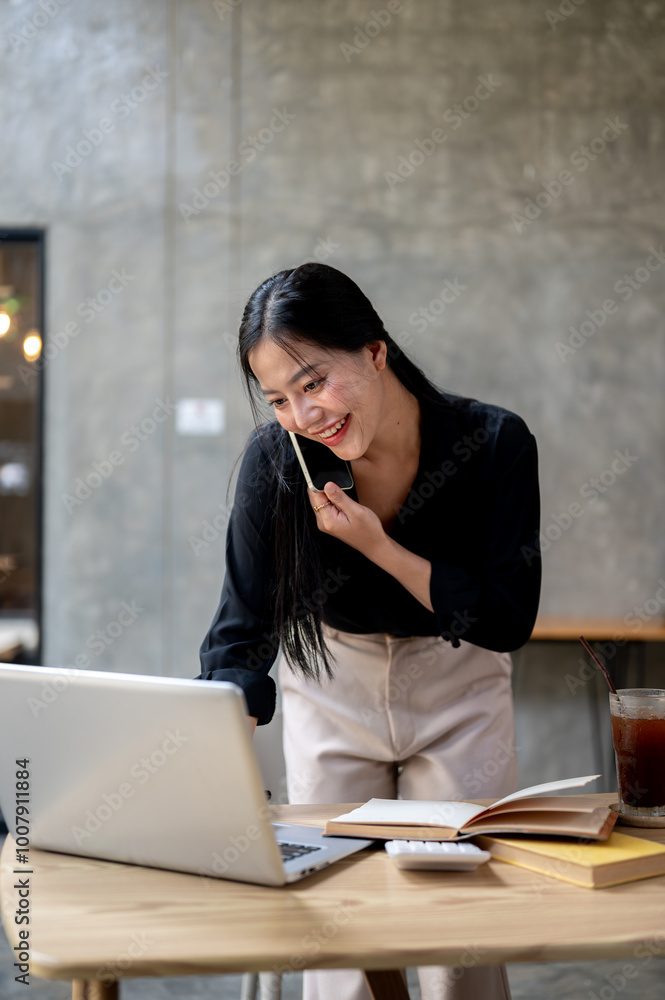 A busy Asian businesswoman is talking on the phone while leaning on her desk and using her computer. A busy Asian businesswoman is talking on the phone while leaning on her desk and using her computer.