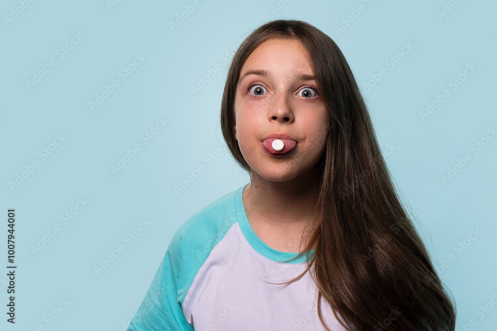 A young girl playfully holds a pill on her tongue, widening her eyes with a humorous expression. A young girl playfully holds a pill on her tongue, widening her eyes with a humorous expression.