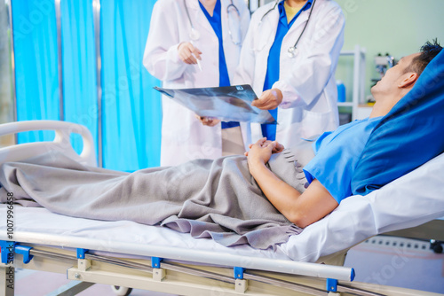 Tableau sur toile A middle-aged man lies bedridden on hospital bed, closely monitored by an old Asian woman doctor and young nurse