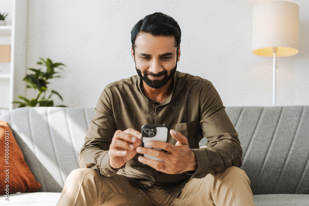 Smiling Indian man sitting on sofa holding mobile phone, using mobile app at home, text message Smiling Indian man sitting on sofa holding mobile phone, using mobile app at home, text message