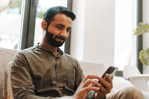 Smiling bearded Indian man holding mobile phone using mobile app, sitting on comfortable sofa