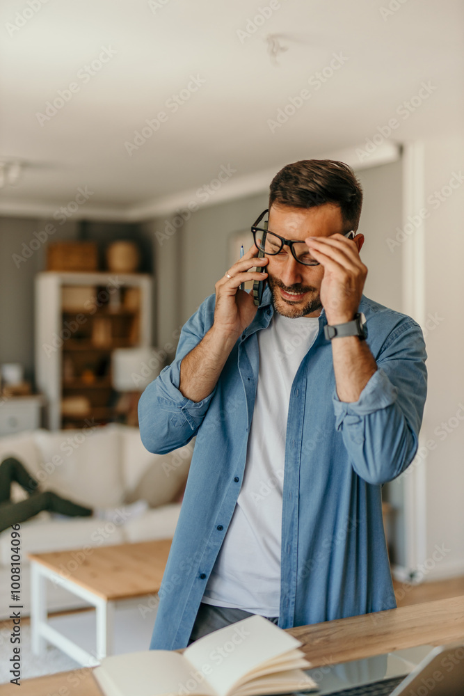 Smiling mid aged man in a denim shirt standing in his living room and talking on the phone during work from home Smiling mid aged man in a denim shirt standing in his living room and talking on the phone during work from home
