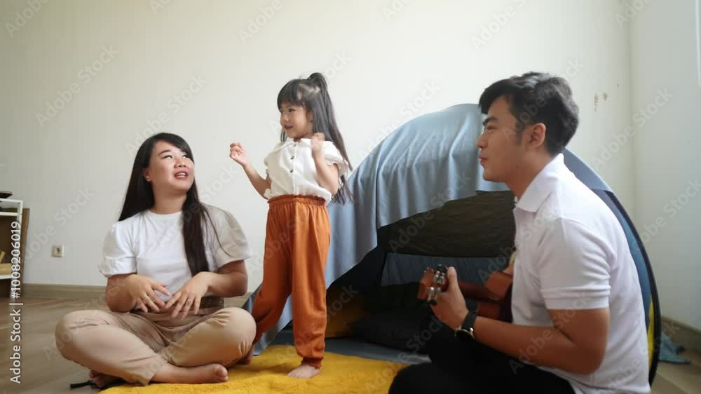 Happy Asian Father Playing Ukulele Guitar While Little Daughter And Mother Singing Together In Front Of Tent At Home
