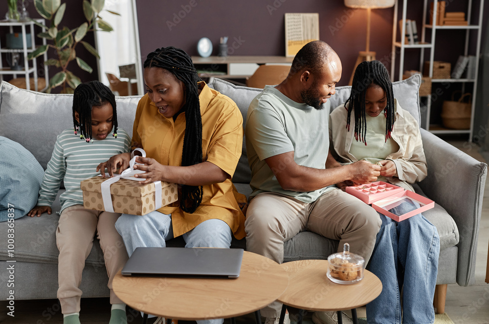 Family enjoying time together on couch while unwrapping gifts and interacting with tablet in cozy living room, displaying connection and happiness Family enjoying time together on couch while unwrapping gifts and interacting with tablet in cozy living room, displaying connection and happiness