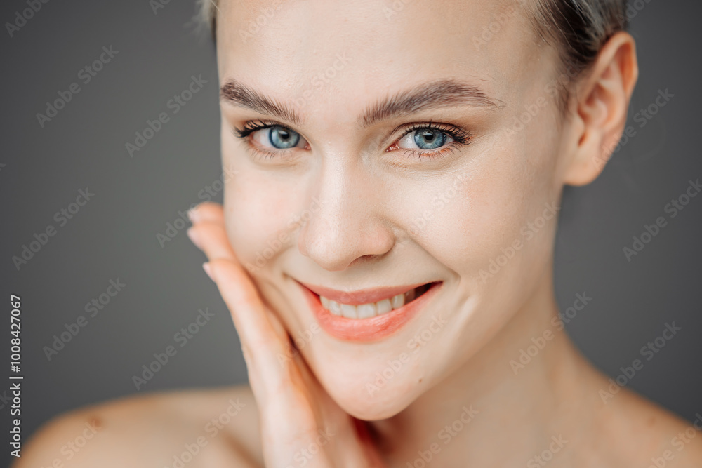 Obraz premium A young woman with radiant skin stands against a dark backdrop, showcasing her natural nude makeup. Her tender touch to her face emphasizes the importance of self-care and beauty.