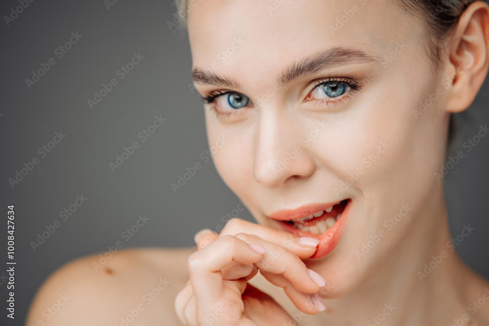 Obraz premium A young woman with radiant skin stands against a dark backdrop, showcasing her natural nude makeup. Her tender touch to her face emphasizes the importance of self-care and beauty.