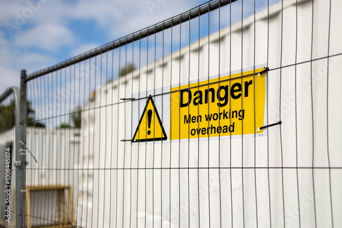 Yellow and black Danger Men Working Overhead triangle warning sign on construction site temporary heras fence panel and white steel storage container photo