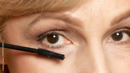 Woman applies mascara with precision, completing her makeup look, emphasizing attention to detail against white studio background. Concept of beauty, wellness, cosmetology, anti-aging products. Ad