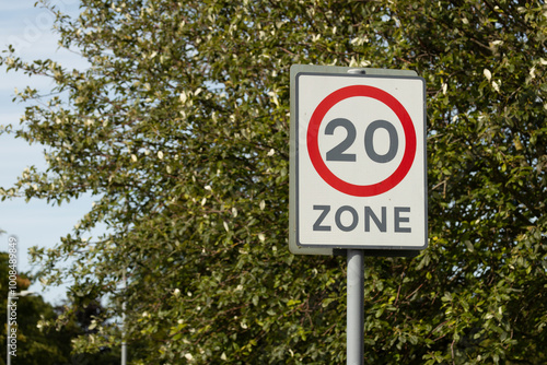 Red circle 20mph zone speed limit road traffic sign in a residential area with trees and leaves