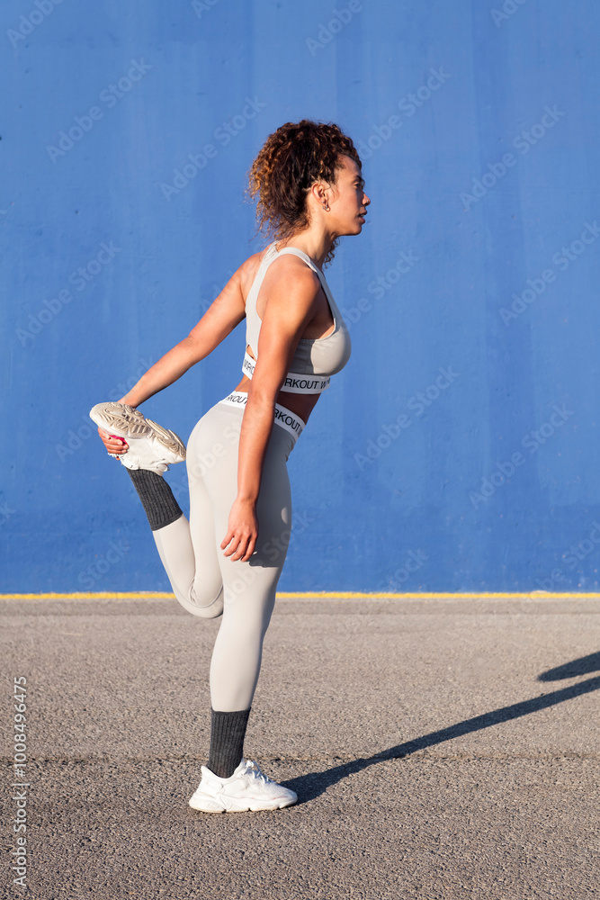 side view of female athlete in sportswear stretching leg during training on asphalt while looking forward on blue background, concept of urban sport and healthy lifestyle side view of female athlete in sportswear stretching leg during training on asphalt while looking forward on blue background, concept of urban sport and healthy lifestyle