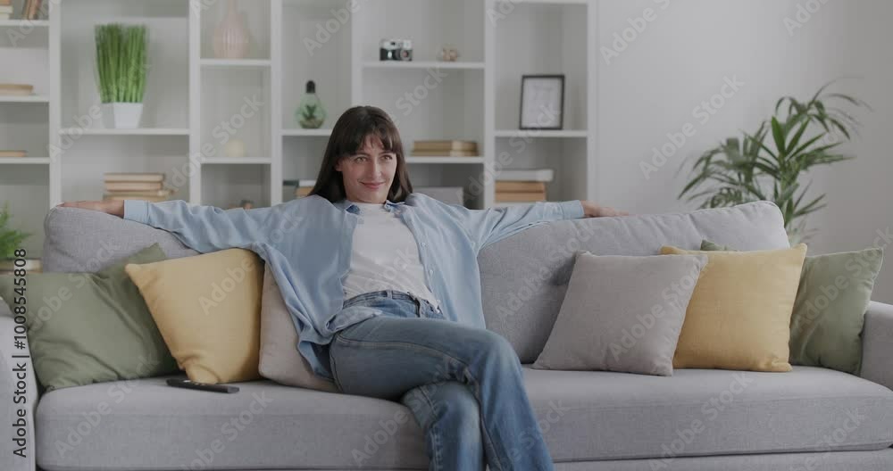 Casually dressed young woman sitting down on a sofa and turns on tv with remote. Casually dressed young woman sitting down on a sofa and turns on tv with remote.
