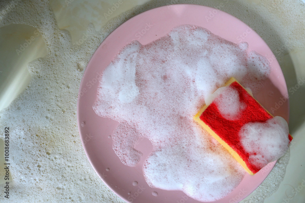 Fototapeta premium Red sponge for washing dishes with a pink plate in soap foam on the kitchen table top view