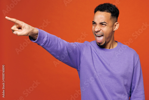 Young sad man of African American ethnicity he wear purple sweatshirt casual clothes point index finger aside scream shout isolated on plain red orange background studio portrait. Lifestyle concept.