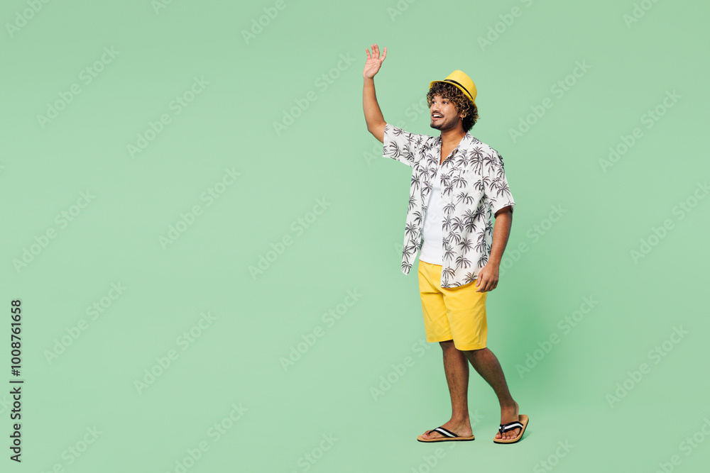 Full body side view young smiling cheerful fun Indian man wear beach shirt hat rest near hotel pool walk go waving hand isolated on plain green color background. Summer vacation sea sun tan concept. Full body side view young smiling cheerful fun Indian man wear beach shirt hat rest near hotel pool walk go waving hand isolated on plain green color background. Summer vacation sea sun tan concept.