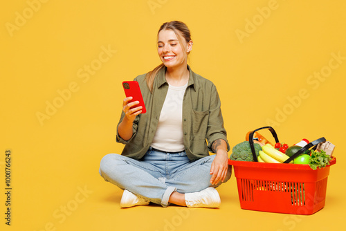 Full body young woman wear casual clothes sit near basket bag for takeaway mock up with food products hold in hand use mobile cell phone isolated on plain yellow background Delivery service from shop photo