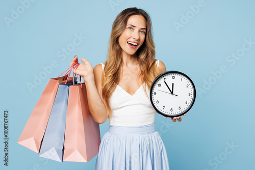 Young fun woman wear casual clothes hold in hand paper package bags after shopping show clock time look camera isolated on plain light pastel blue cyan background. Black Friday sale buy day concept.