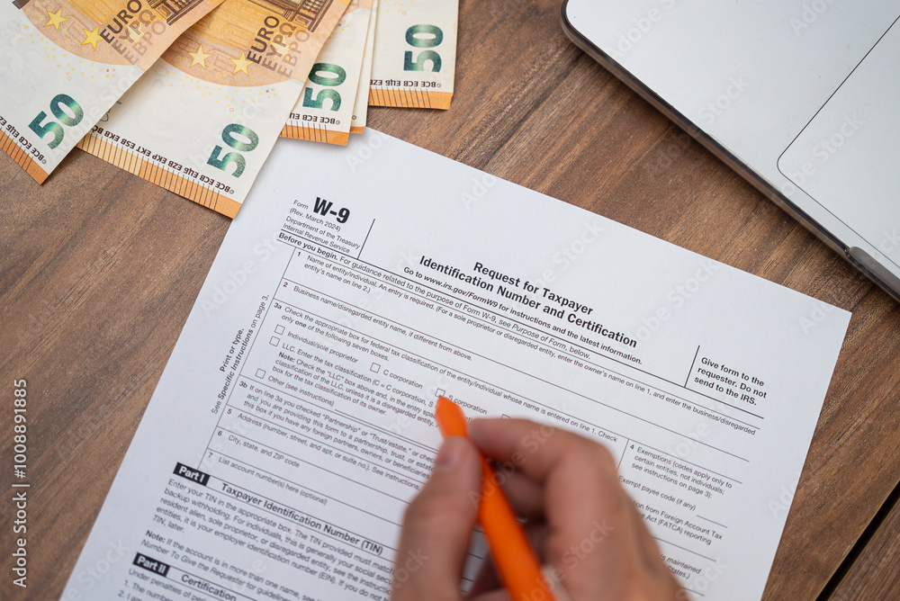 Obraz premium Hand filling out a W-9 tax form with euros and a laptop on a wooden desk. Financial preparation and tax season concept, suitable for accounting and international finance themes 