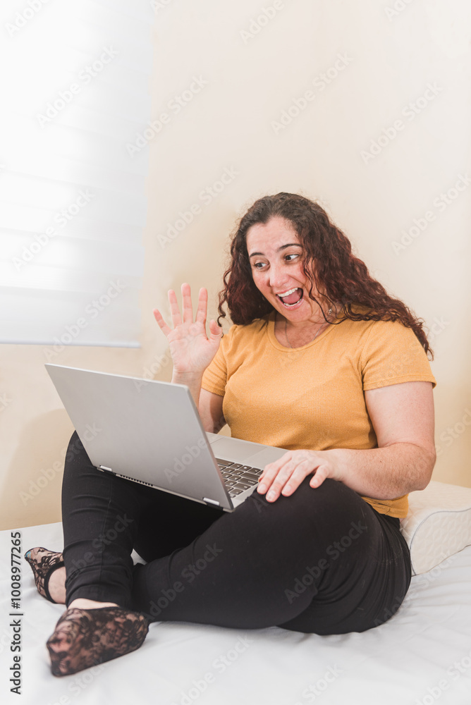 Smiling woman waving during a video call on her laptop, seated casually on a bed. Engaging in remote communication from home. Smiling woman waving during a video call on her laptop, seated casually on a bed. Engaging in remote communication from home.