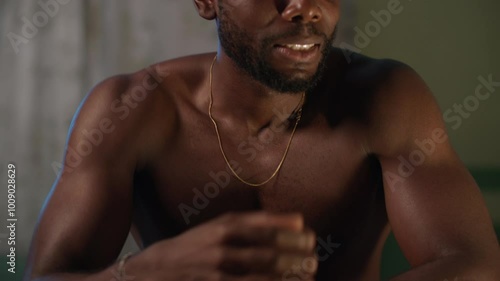 Worried and anxious young shirtless African man sitting on his bed, having hand tremors at night.