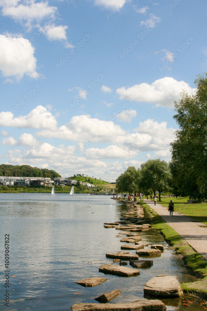 Fototapeta premium Phoenix See, Dortmund, Germany. Sunny summer day with blue skies, boats in the lake, people walking, swans in the water and hill in a background. Vertical