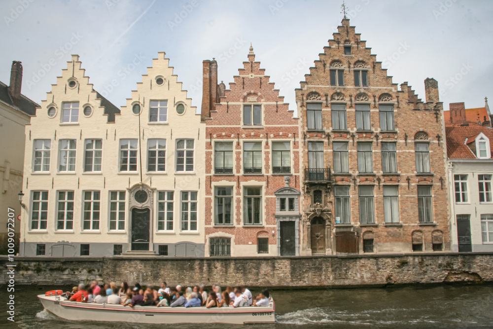 Fototapeta premium A boat with tourists passing tall historic residential buildings next to a canal in Bruges, Belgium