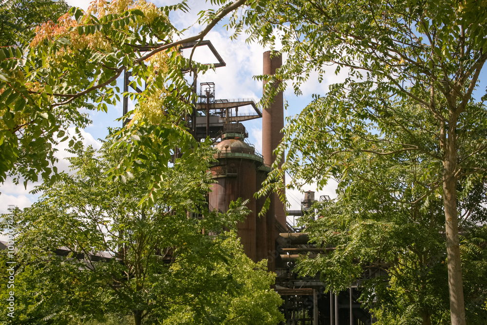 Germany, North Rhine-Westphalia, Dortmund-Hoerde, Phoenix West, abandoned blast furnace steel factory behind green summer tree leafs. Germany, North Rhine-Westphalia, Dortmund-Hoerde, Phoenix West, abandoned blast furnace steel factory behind green summer tree leafs.
