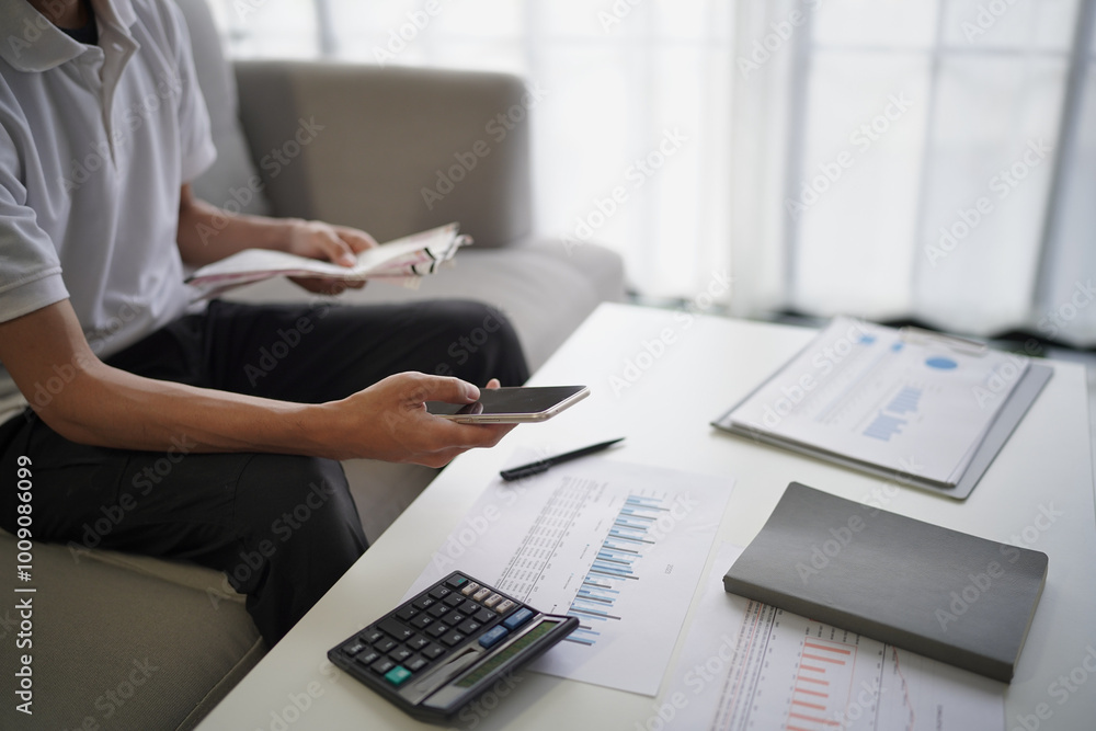 Man checking bills balances Invoices and make payment by online banking. there are calculator and document on table. He sitting on sofa in living room at home Man checking bills balances Invoices and make payment by online banking. there are calculator and document on table. He sitting on sofa in living room at home