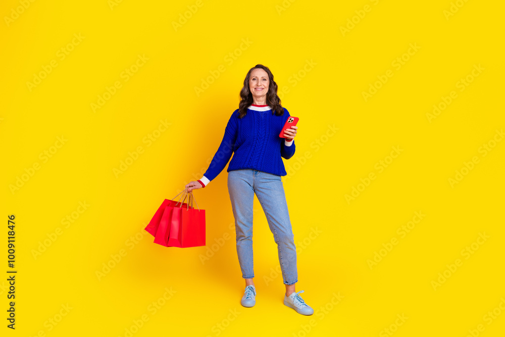 Full length photo of lovely pensioner lady device shopping bags dressed stylish blue knitted garment isolated on yellow color background Full length photo of lovely pensioner lady device shopping bags dressed stylish blue knitted garment isolated on yellow color background